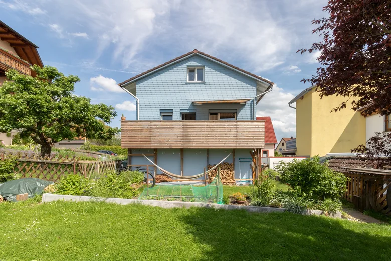 Blau verschindeltes Einfamilienhaus mit Holzbalkon, davor ein Garten mit Obstbaum.